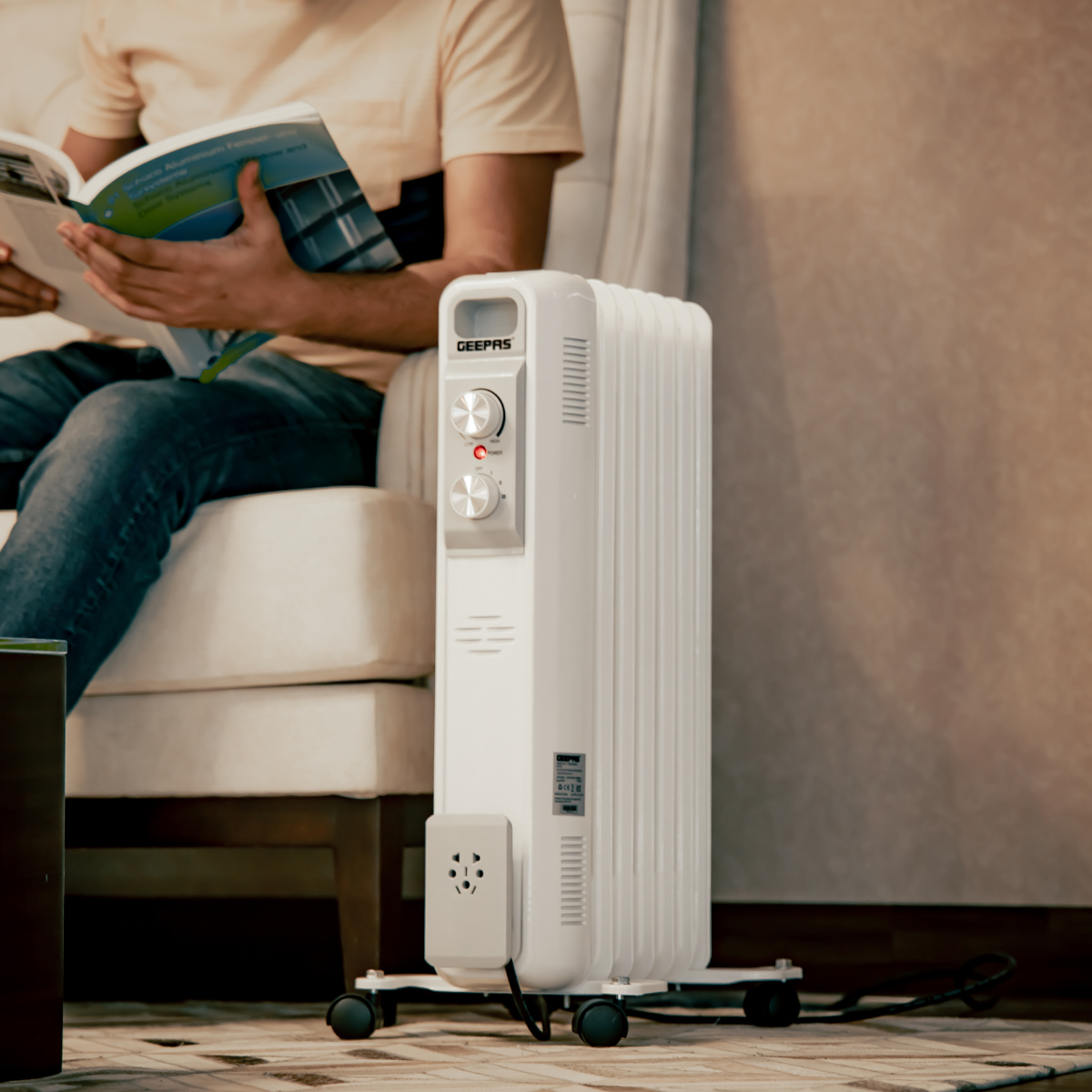 Image of the electric 7 fin oil filled radiator on a living room floor with a person sitting behind it reading a magazine