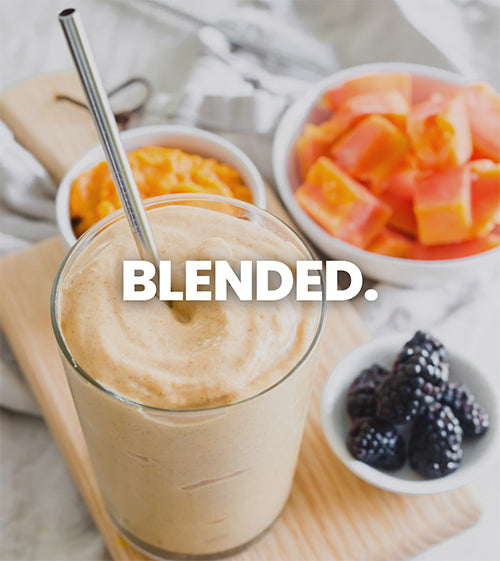 Image shows a few different fruits in white bowls ready to be blended into a smoothie, there is a glass full to the top with a smoothie with ice