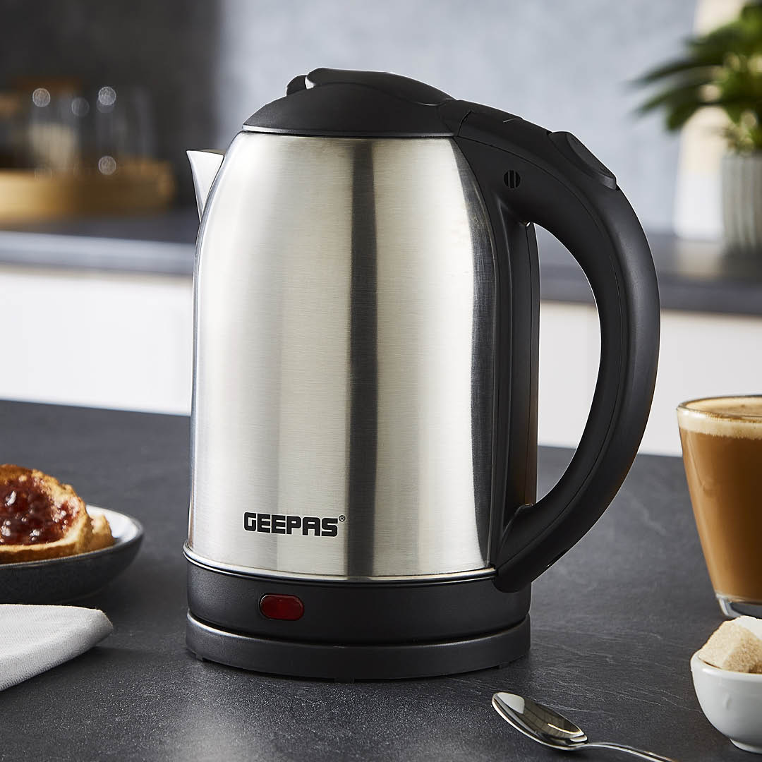 The image shows off the geepas stainless steel kettle on top of a kitchen countertop with a cup of coffee and a piece of toast with jam besides it.