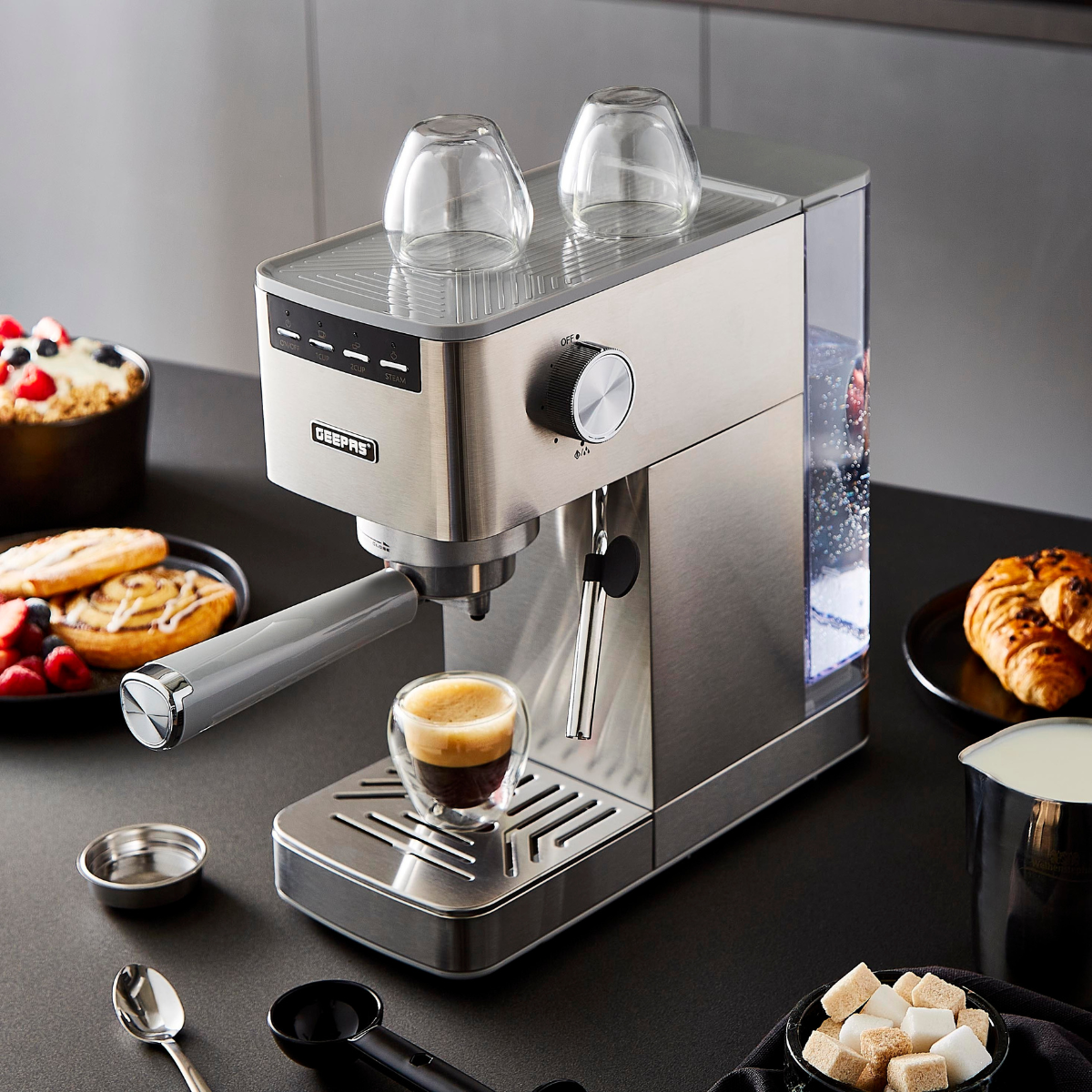 The image shows the long espresso coffee machine on top of a kitchen countertop with several baked goods around it.