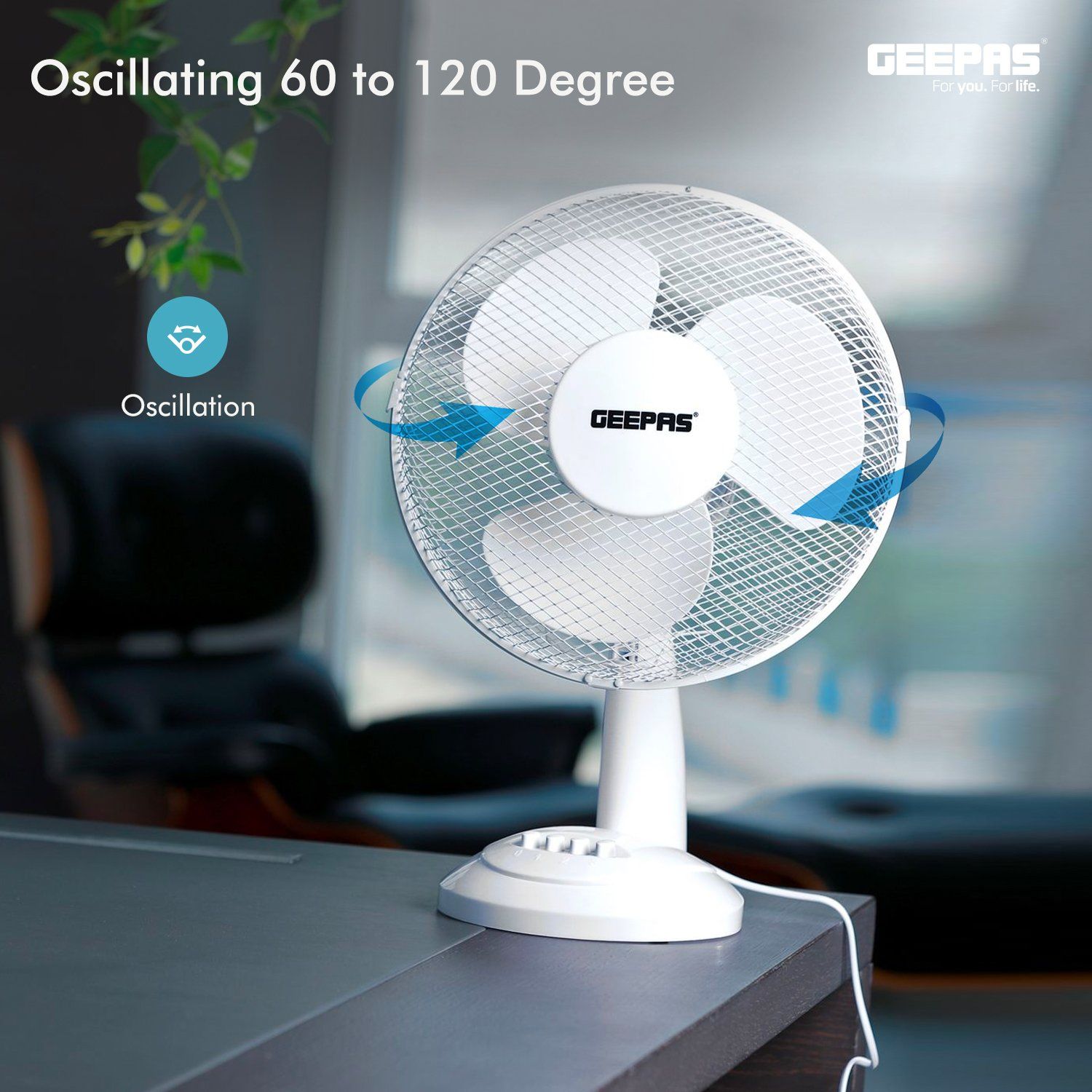 There is a white cooling fan on top of a grey wooden office desk with office furniture and a panoramic view in the background.