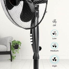 The image shows off a close up photograph of the black pedestal fan it shows what the four different buttons do, the first button on the top shows to power the fan off, the next one down shows low speed, the next one down shows medium speed setting, the last one down shows the highest speed. There are also some living room decorations in the background of the image such as plants and a sofa.